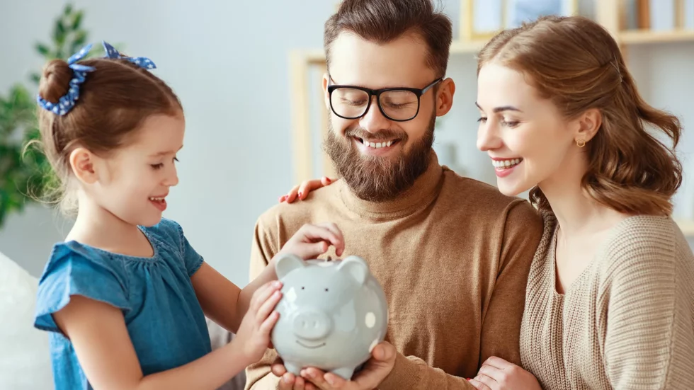 imagem de uma familia com um cofre de dinheiro nas mãos estimulando a filha pequena a guardar moedas
