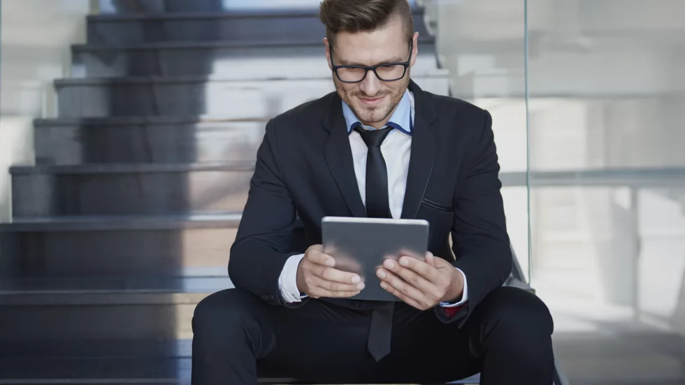 Imagem de um empresário sentado em uma escada analisando seriamente a tela do seu tablet